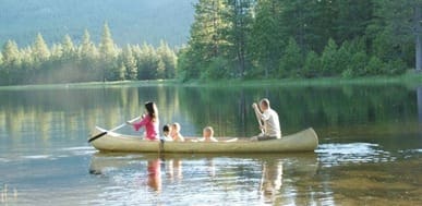 Family canoeing on a calm lake surrounded by evergreen trees and mountains.