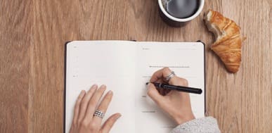 Open planner, coffee, and croissant on a wooden desk for morning routine planning.