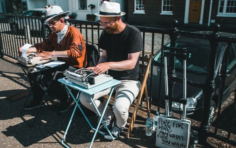 Two poets for hire typing on vintage typewriters outdoors; sign says "Poet for Hire, Pay Whatever You Want
