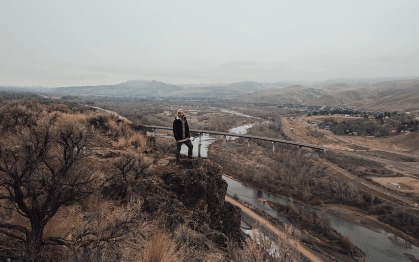 Alex Cwalinski stands on a cliff overlooking a river and bridge, with mountains in the background.