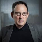 Man with glasses in a grey blazer, looking at the camera with a serious expression