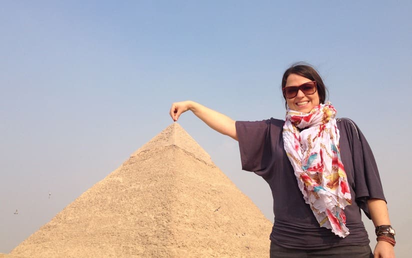 Woman playfully touching the top of a pyramid in Giza, Egypt, wearing sunglasses and a colorful scarf.