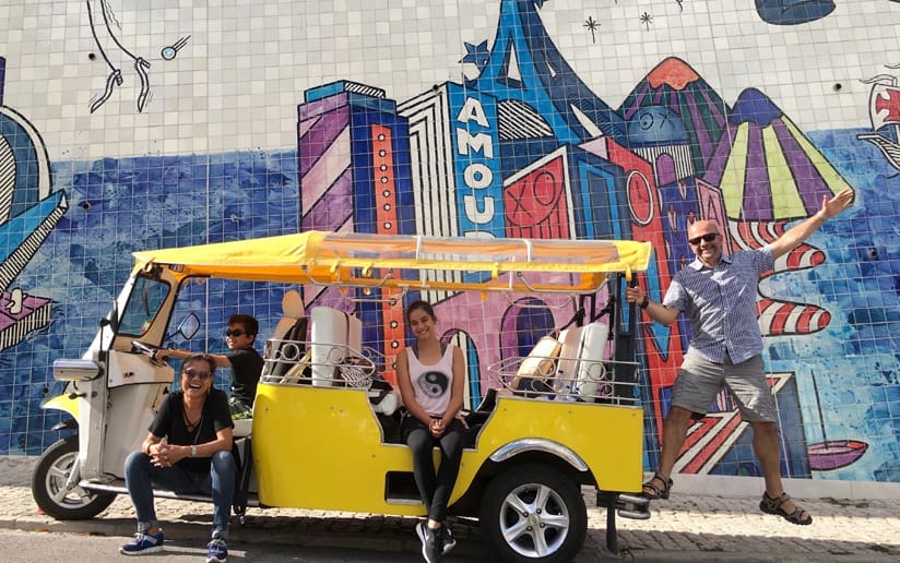 Family posing with a yellow tuk-tuk in front of a colorful mural. Fun vacation photo.
