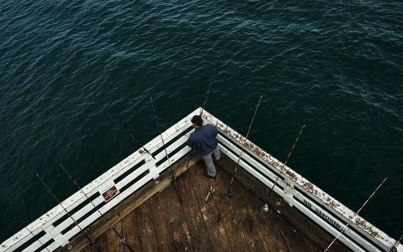 Fisherman on pier surrounded by fishing rods, deep blue ocean water.