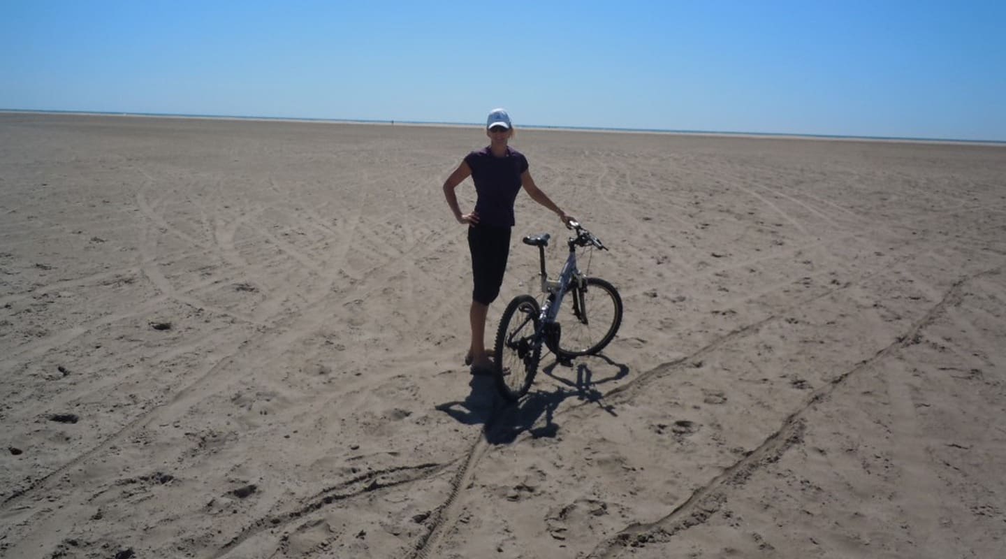 Woman with bicycle on sandy beach under a clear blue sky.