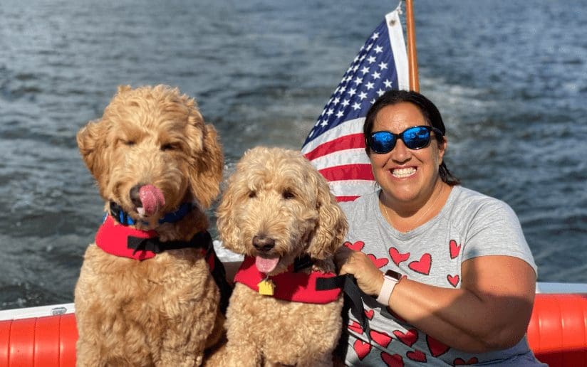 Michelle Harden with two Goldendoodle dogs wearing life jackets on a boat with an American flag.