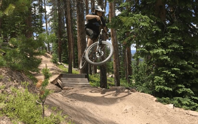 Mountain biker catching air off a wooden jump in a forest setting.