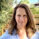 Woman with long brown hair smiling outdoors, wearing a blue striped shirt.