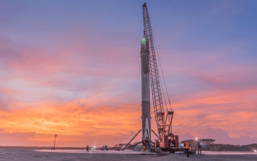 SpaceX rocket being prepared for launch with a crane against a colorful sunset sky.