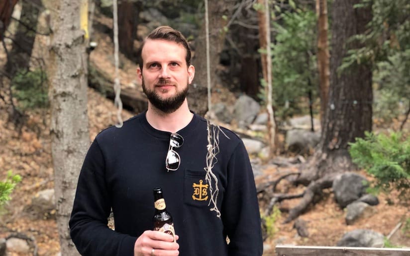 Man with beard holding a beer outdoors in a wooded area