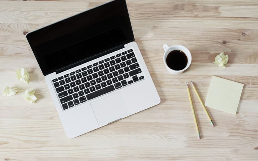 Laptop, coffee, pencils, and crumpled paper on a wooden desk, ideal for writing.