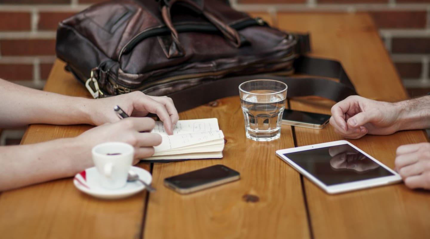 Meeting with consultant: hands writing in notebook, coffee, tablet, and phone on wooden table.