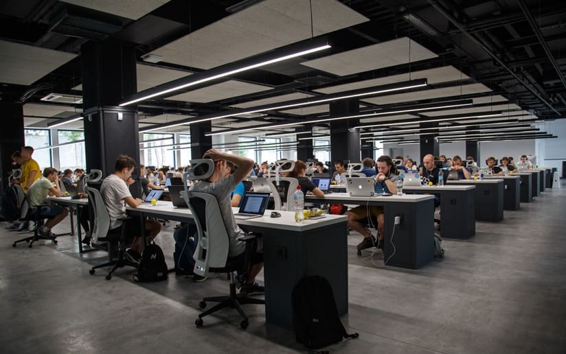 Busy open-plan office with rows of employees working at desks with laptops.