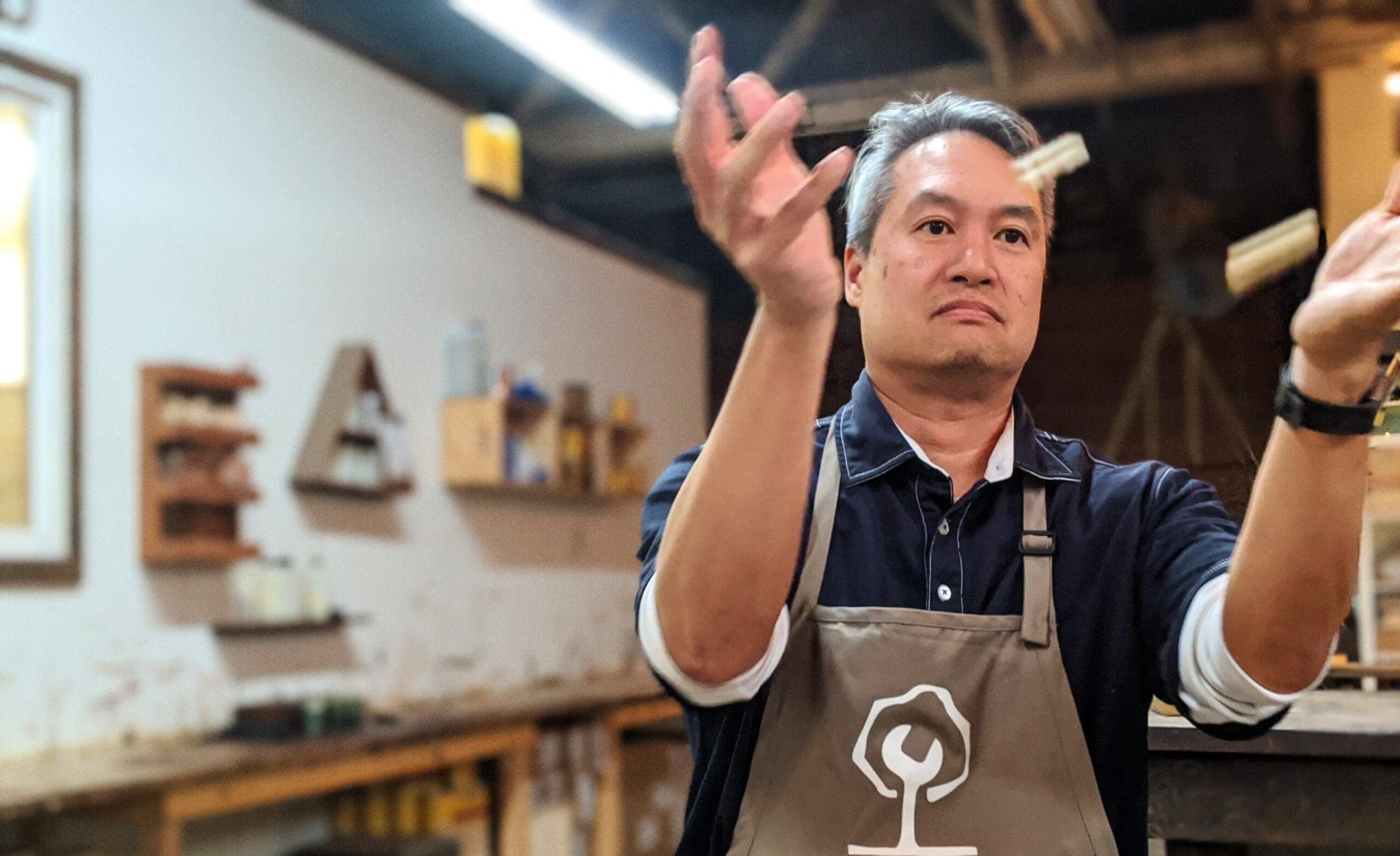 Man tossing wooden objects in workshop, wearing apron. Background shows tools and work area.