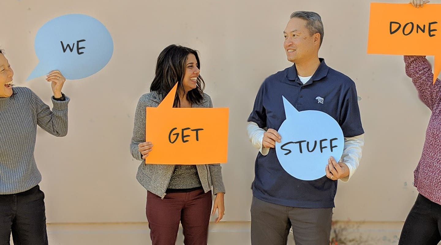 Four people holding speech bubbles saying We Get Stuff Done in a collaborative setting.