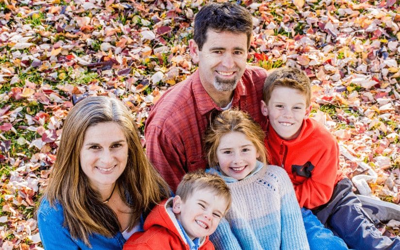Family portrait with two parents and three kids smiling in a pile of autumn leaves.