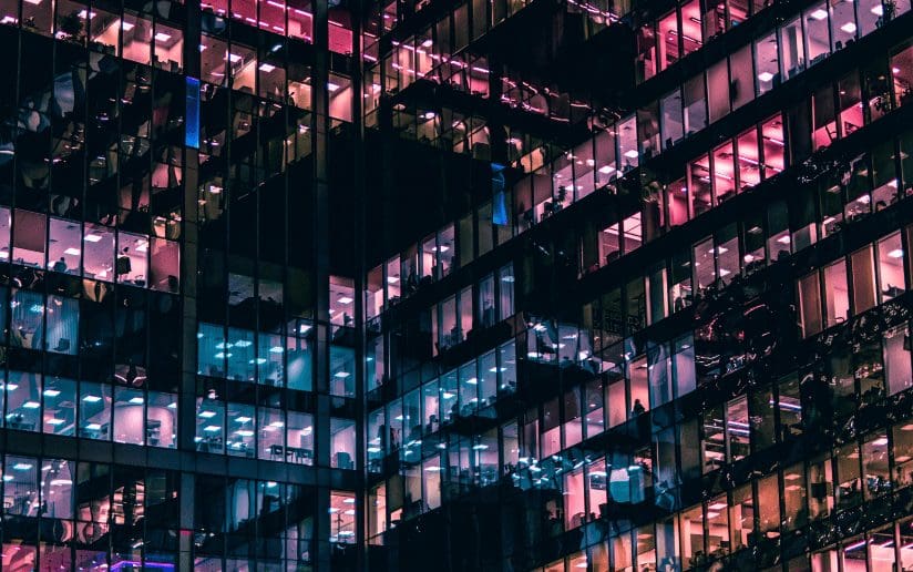 Office building windows lit up at night, reflecting pink and blue hues.