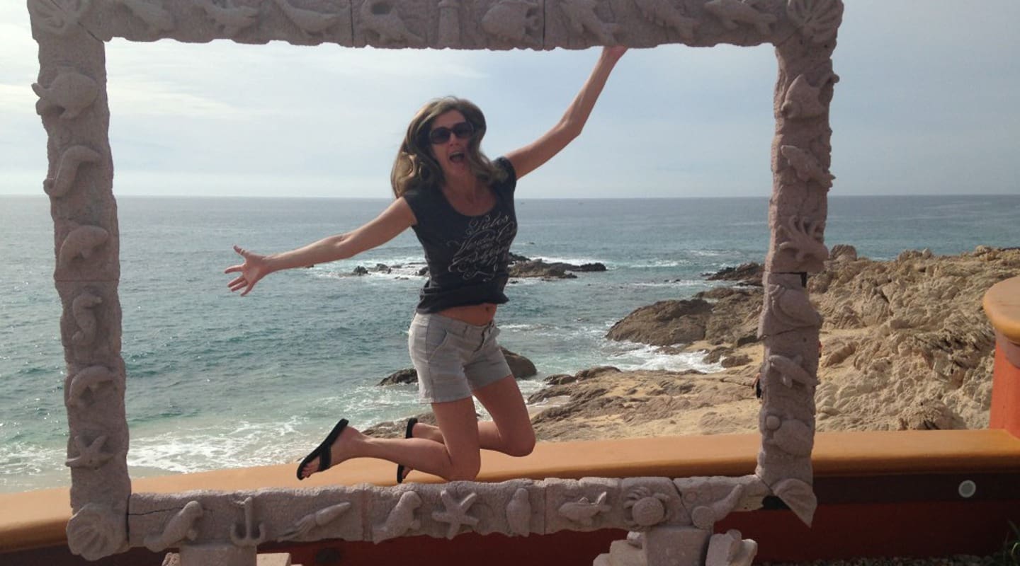 Jen Roop jumping for joy near the ocean, wearing sunglasses and shorts.