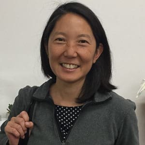 Woman with dark hair smiles, wearing a grey jacket and polka dot shirt.