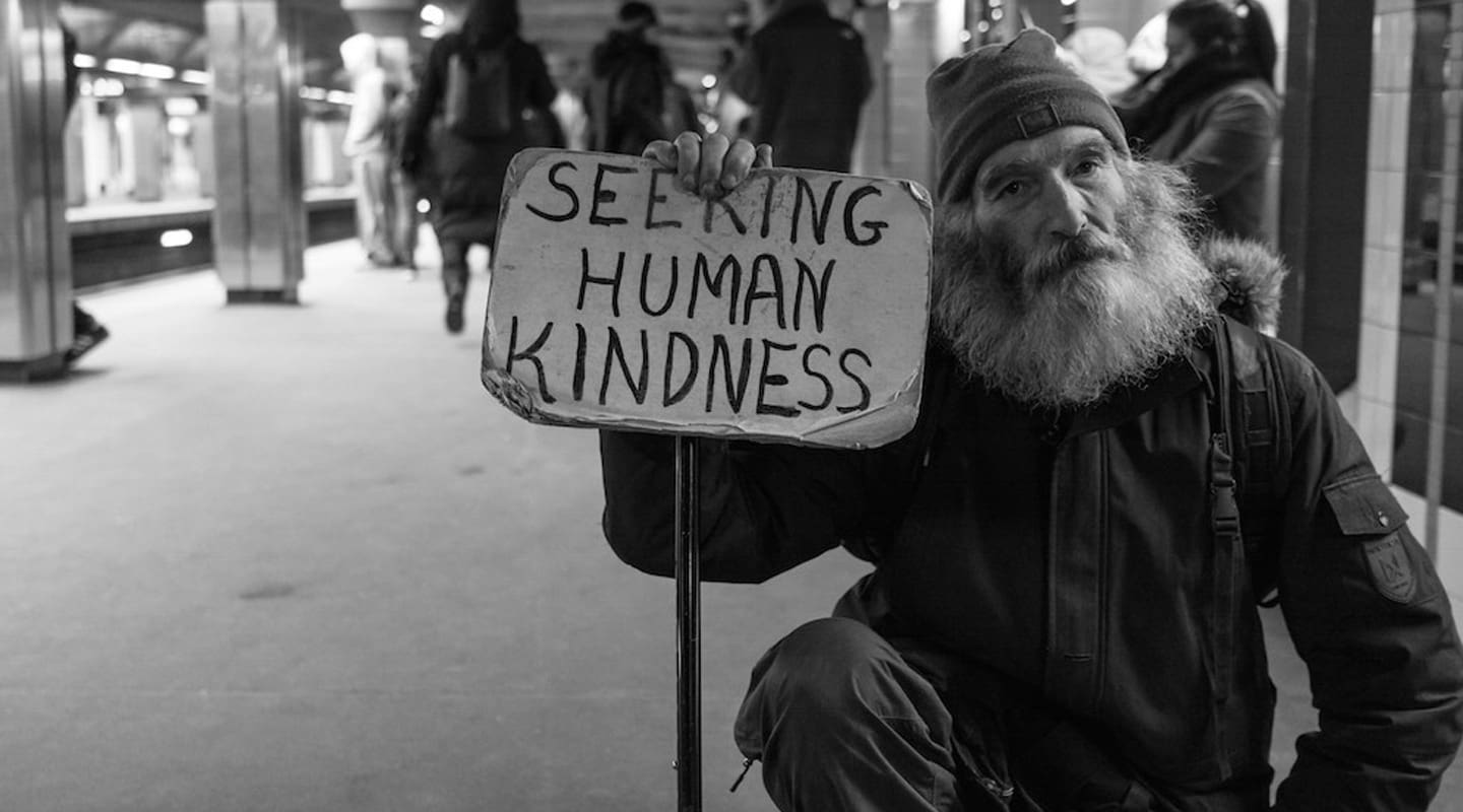 Man holding Seeking Human Kindness sign in a public space; related to social good.