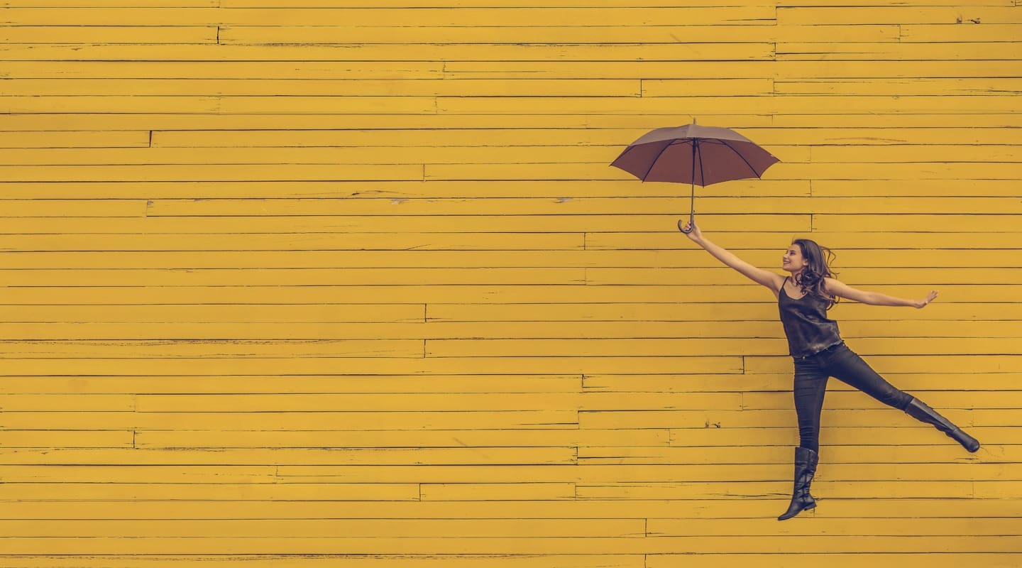 Woman levitating with umbrella against a yellow wall, symbolizing stress reduction and work-life balance.