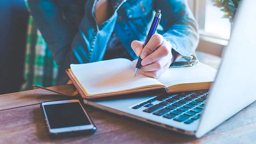 Person writing in notebook next to laptop and smartphone, focusing on note-taking and productivity.