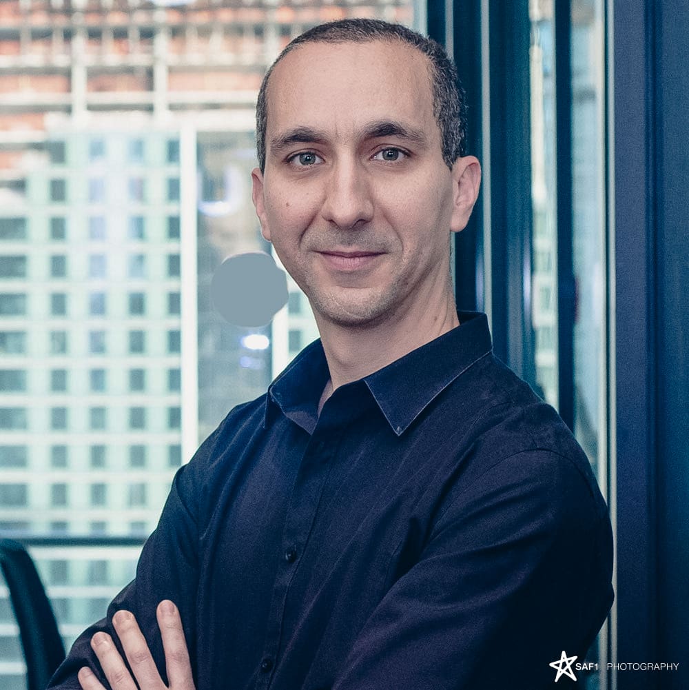 Man in a black shirt with arms crossed, smiling in a modern office setting. Leadership.