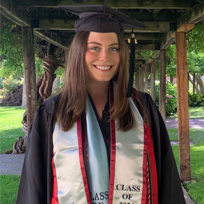 Alexa Pritchard, a young woman wearing a black graduation cap and gown, standing outdoors beneath a wooden pergola surrounded by greenery. She’s smiling and wearing a light blue graduation stole with red trim that reads Class of 2023.
