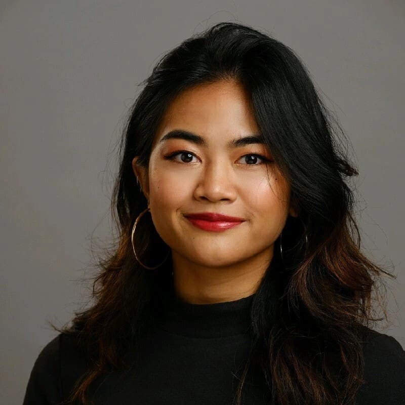 Bianca Camacho, a woman with long, dark hair wearing hoop earrings and a black turtleneck top. She is smiling softly against a neutral gray background in a professional headshot.