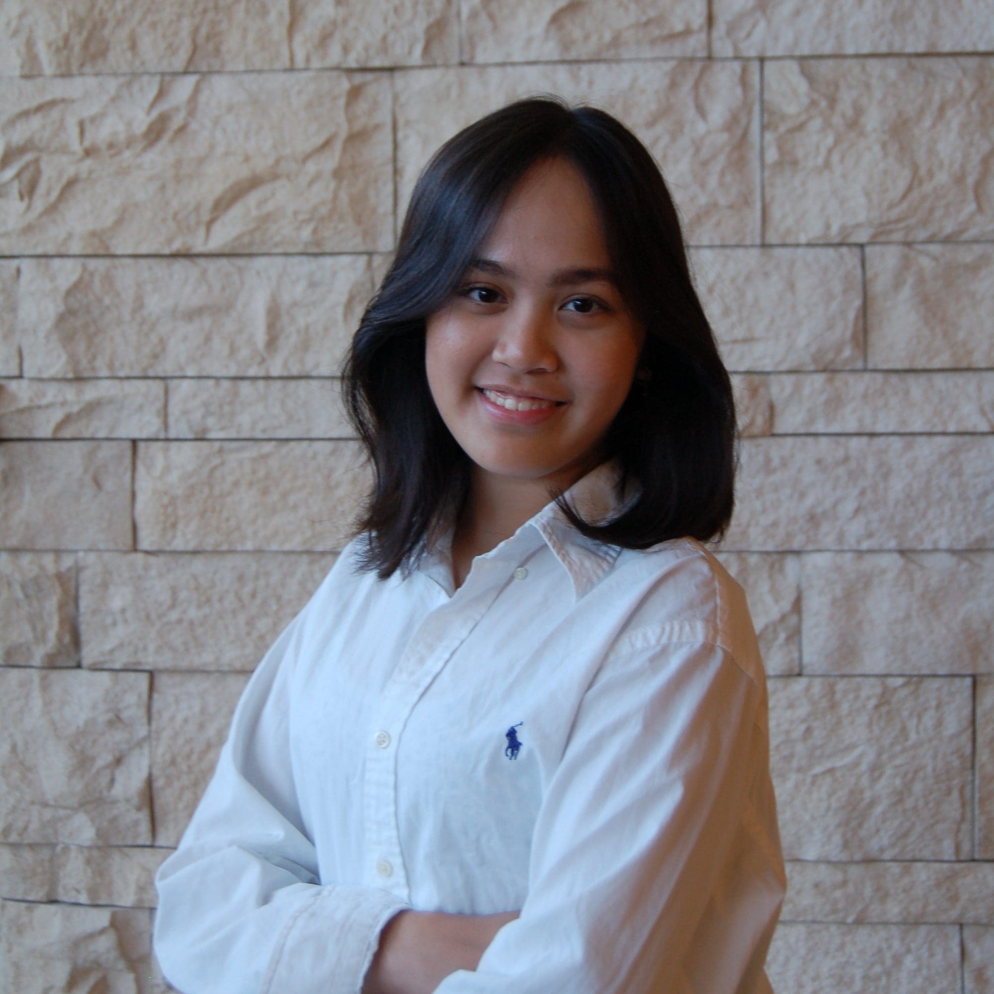 Claudia Regiarto, a woman with shoulder-length dark hair standing in front of a beige stone wall. She is smiling, dressed in a white button-down shirt with the sleeves casually rolled up.