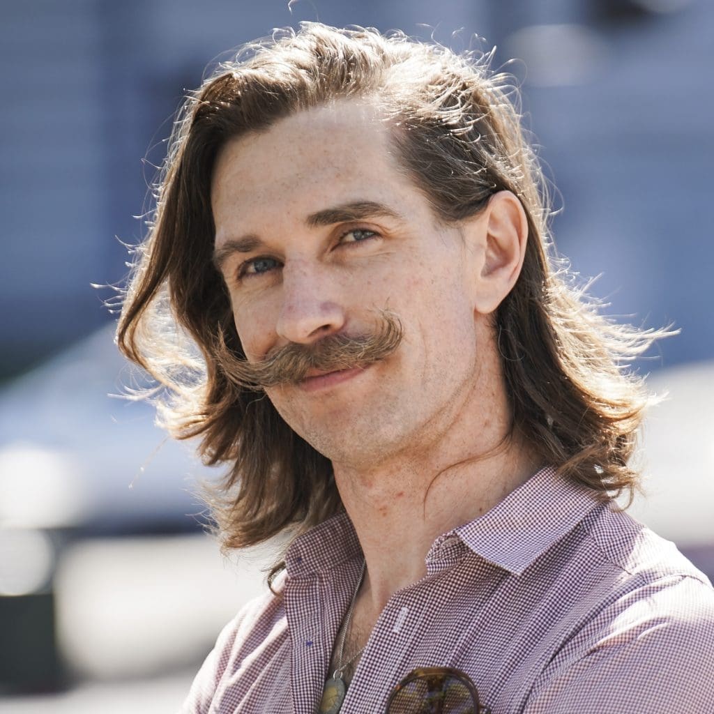 Portrait of Justin Liszanckie outdoors, wearing a button-up shirt with long hair and a distinctive handlebar mustache.