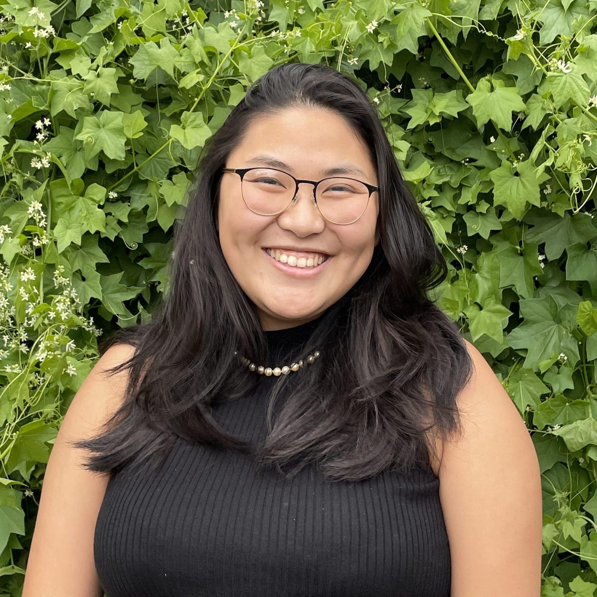 Brittney Kitaura, a woman with long, dark hair wearing glasses, a black sleeveless top, and a pearl necklace. She’s standing in front of a vibrant green wall of ivy, smiling warmly at the camera.